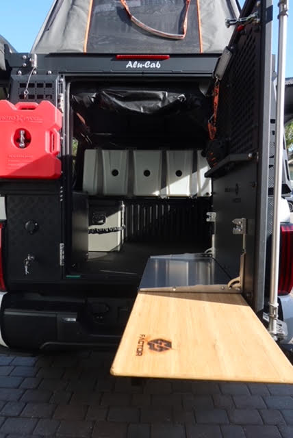 Rear of the camper open with the GP Factor foldaway bamboo kitchen table deployed off the tailgate, showing the Alu-Cab badge, side storage box, Rotopax mount and the fresh water tanks inside the bed — photo 7