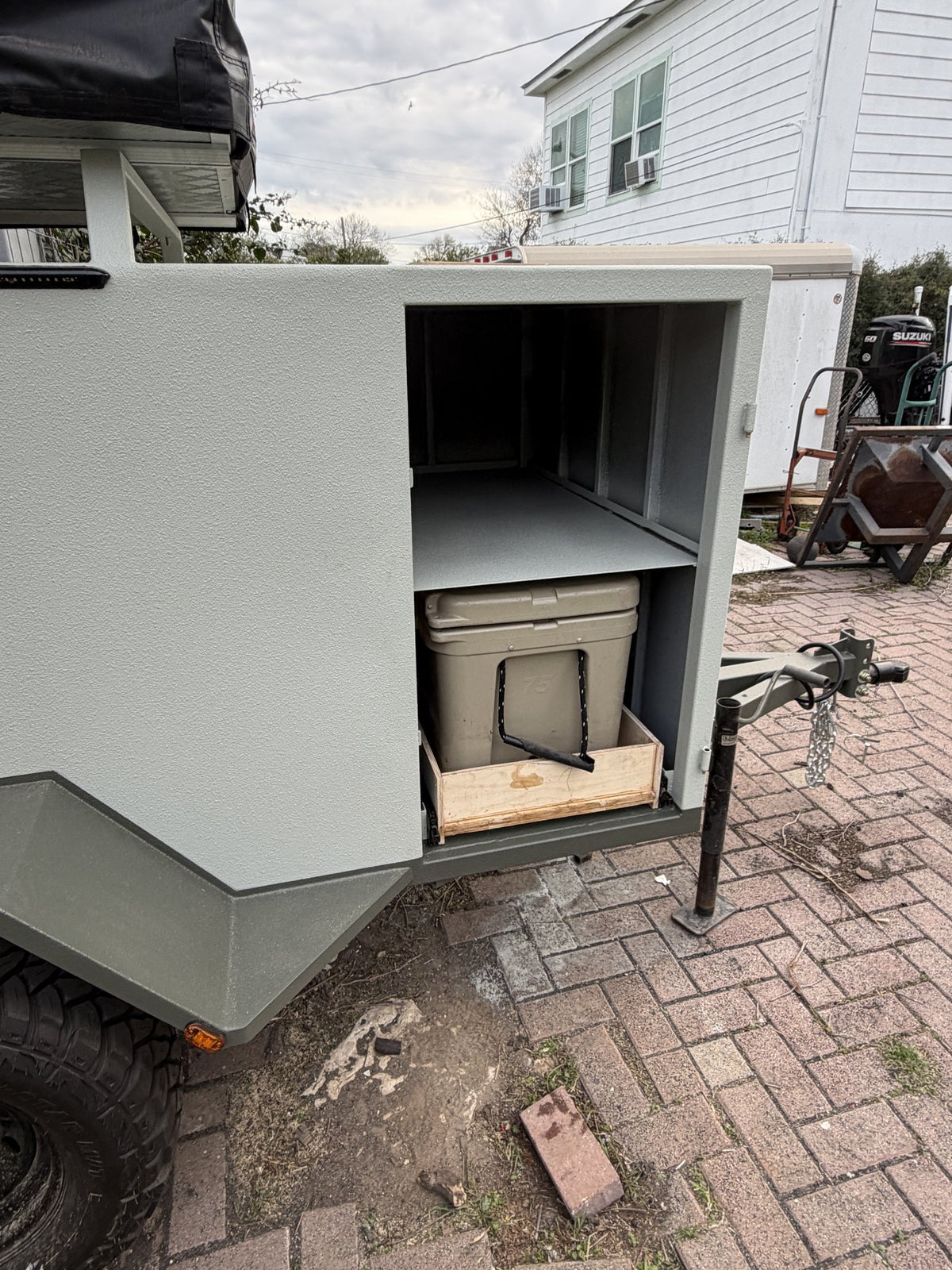 Side hatch open on the passenger side showing the upper shelf and the lower slide-out drawer with a tan 75-quart ice chest strapped in place, trailer tongue and jack visible at right — photo 5