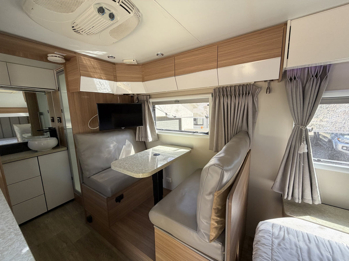 Interior looking forward from the bedroom: grey leatherette dinette with pedestal table under curtained windows, wall-mounted flat-screen TV, overhead cabinetry, roof air-conditioning unit and the bathroom vanity beyond — photo 6