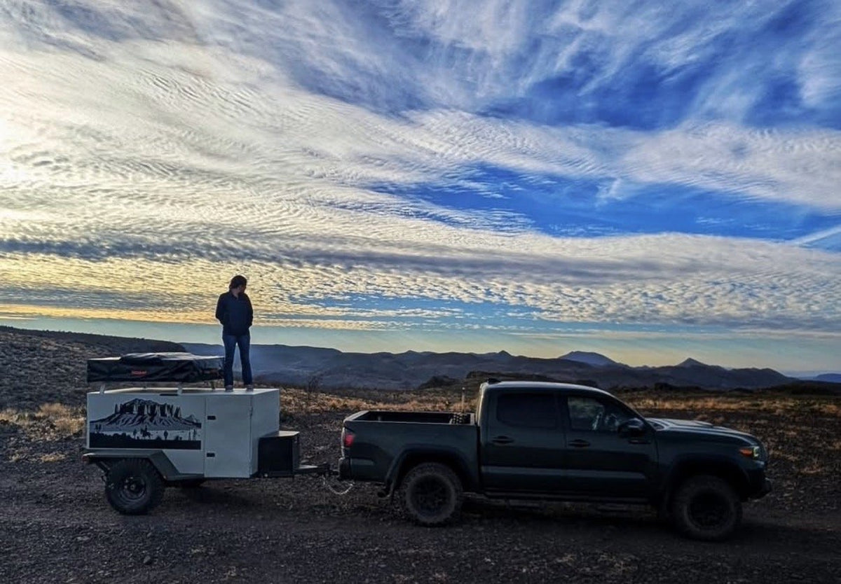 The trailer hitched behind a Toyota Tacoma at a high desert overlook at sunset, a person standing on the trailer roof beside the packed rooftop tent, mountains and streaked clouds in the distance — photo 6
