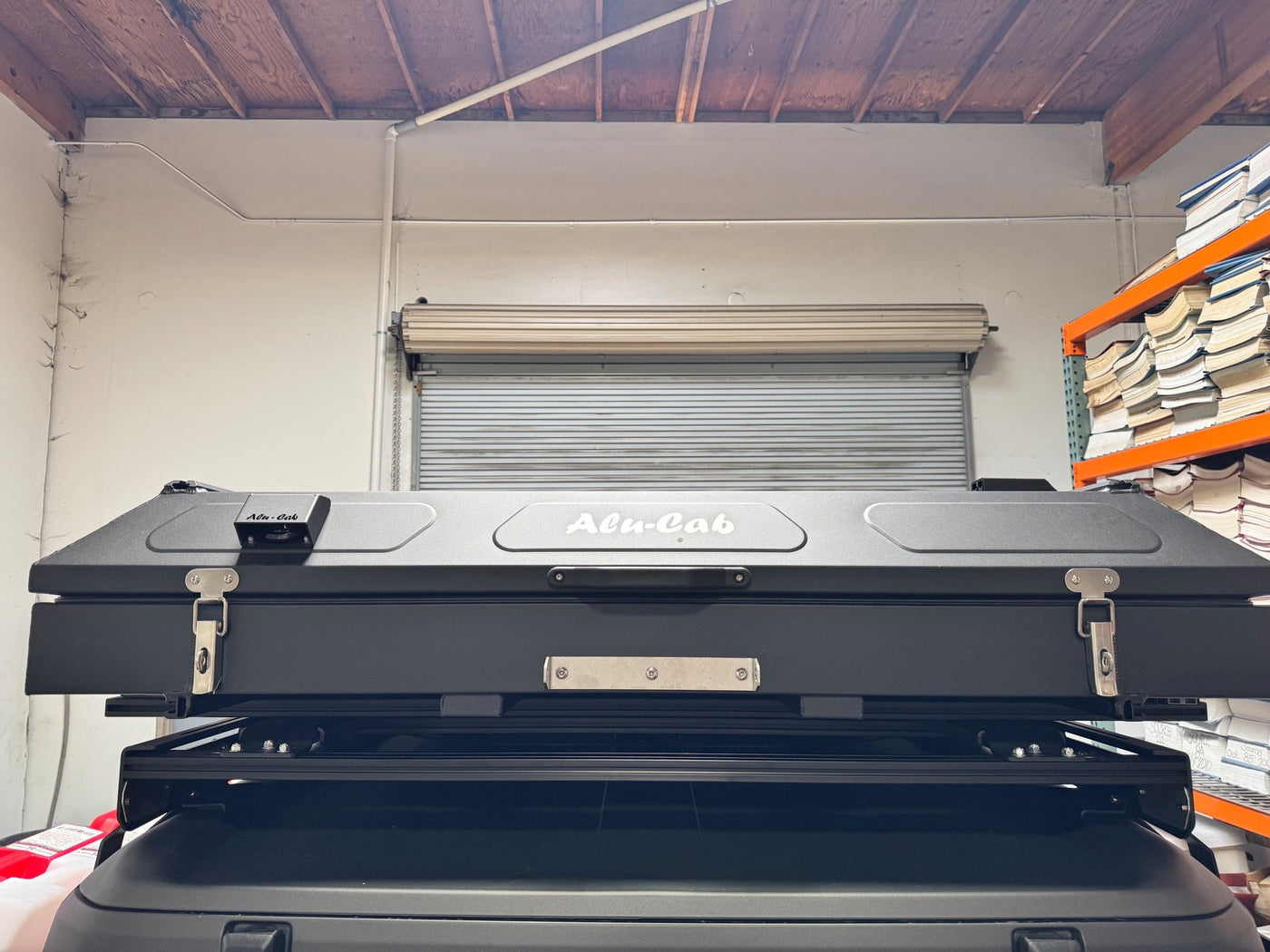 The Alu-Cab Gen 3-R rooftop tent closed and latched on its roof rack, viewed straight on, showing the black aluminum shell with the Alu-Cab logo, side latches and mounting brackets — photo 3