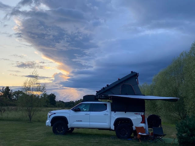 The rig at a grass campsite at sunset with the 270-degree shadow awning extended over camp chairs, camper roof raised against a cloudy evening sky — photo 10