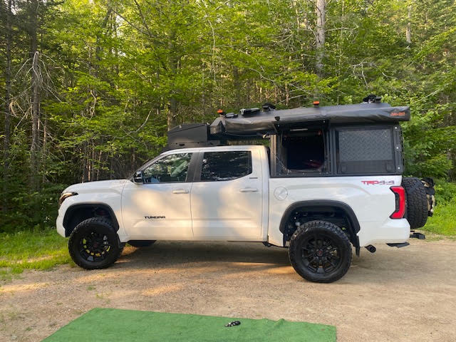Driver-side profile at a wooded campsite with the Alu-Cab Cabin roof popped up, showing the raised tent section, gull-wing side door open and the Sherpa roof rack over the cab — photo 3