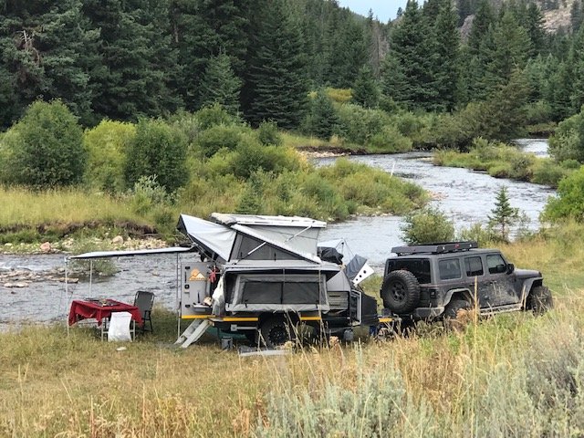 Camp set up on a river bank with the Sundowner's hard-shell rooftop tent raised, side awning extended, camp chairs and table out, and the Jeep parked alongside — photo 6