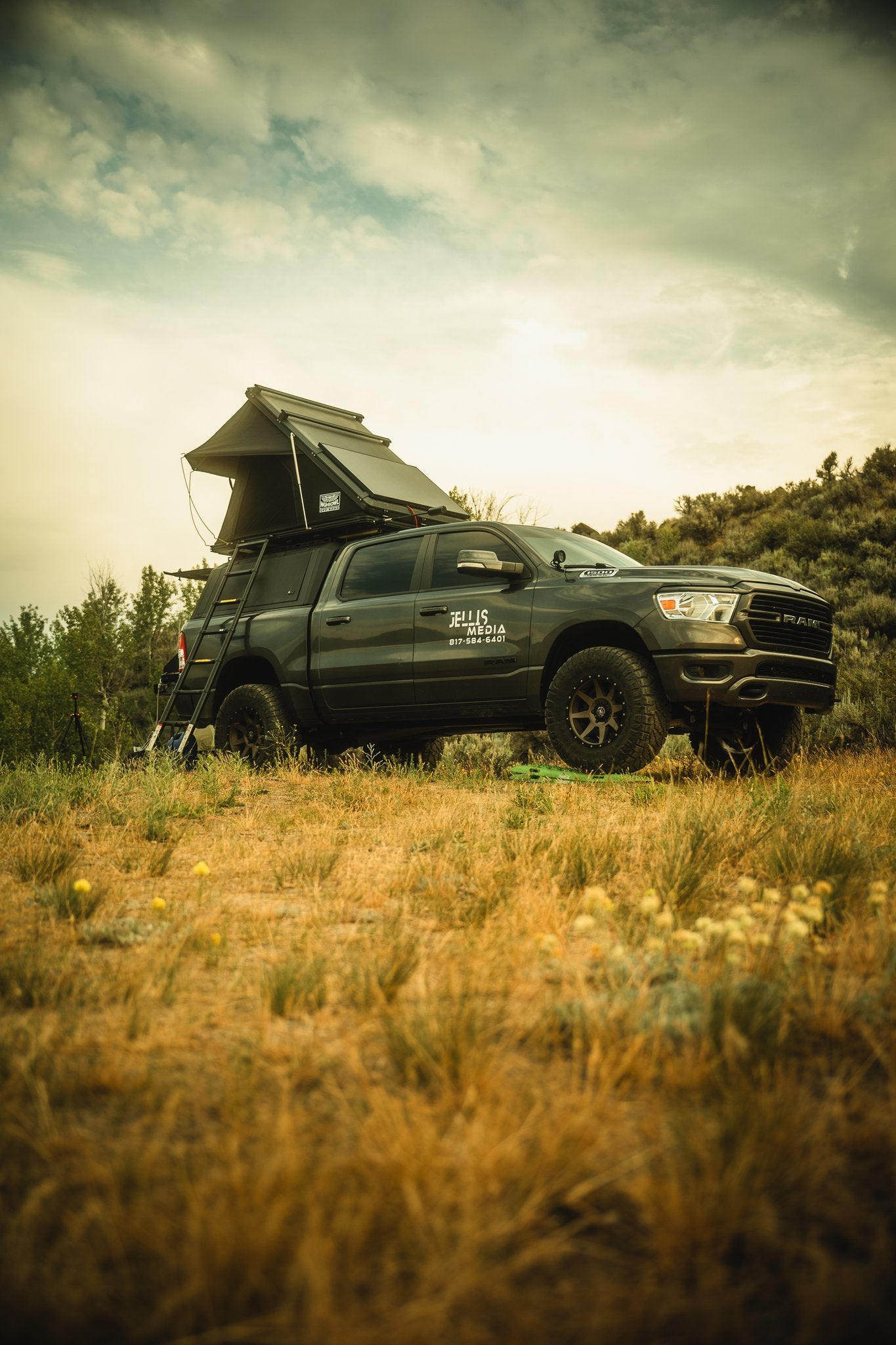 Driver-side view in tall grass under storm clouds, rooftop tent deployed on the camper shell with the ladder extended, showing the shell's side window and the crossbars running across the shell roof — photo 3
