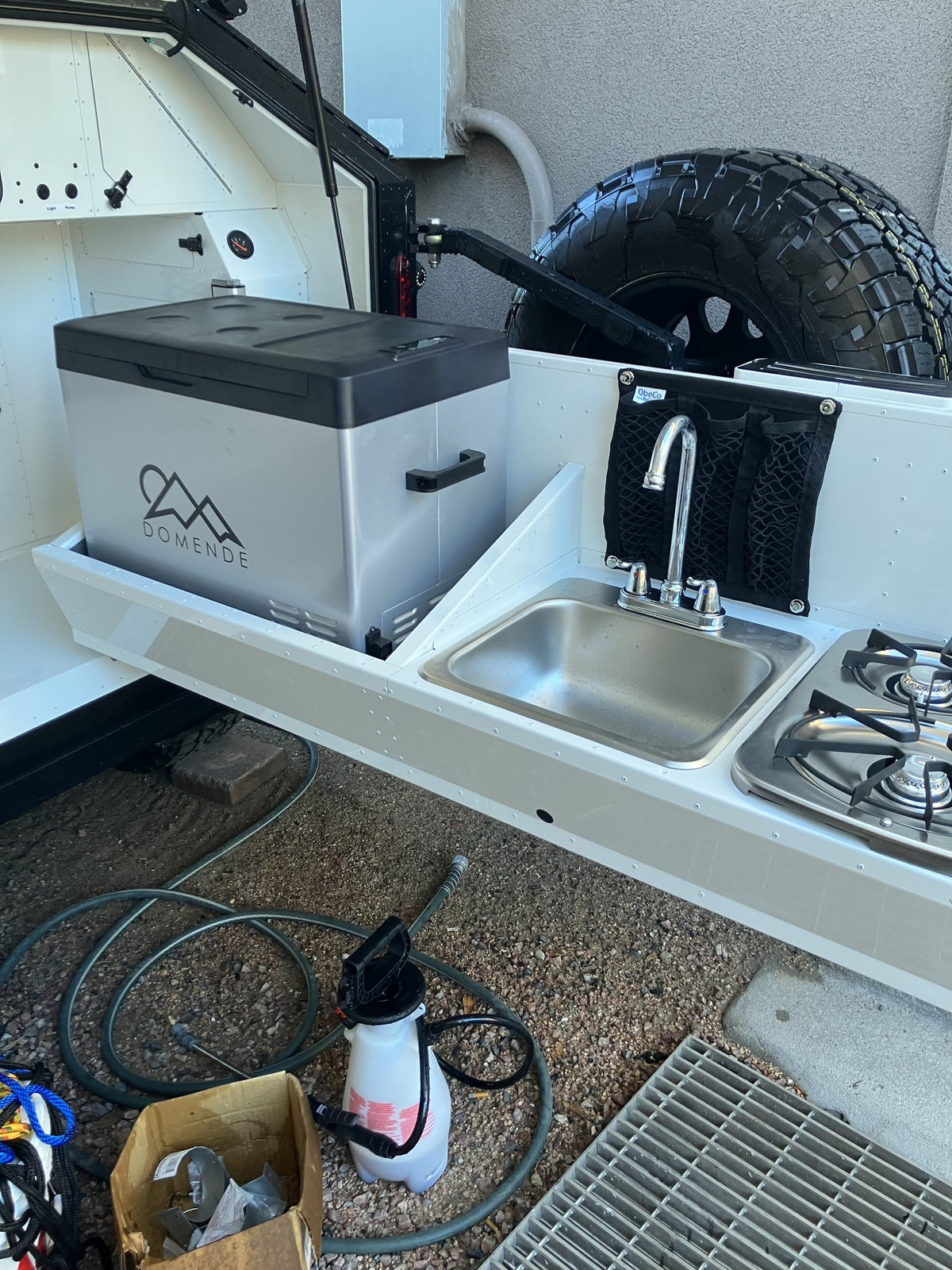 Slide-out galley of the 2023 Mammoth Overland ELE: stainless steel sink with a chrome faucet, a two-burner propane cooktop, a DOMENDE portable fridge/freezer on the pull-out tray, a mesh utensil pocket, and a full-size spare tire on the rear carrier behind — photo 5