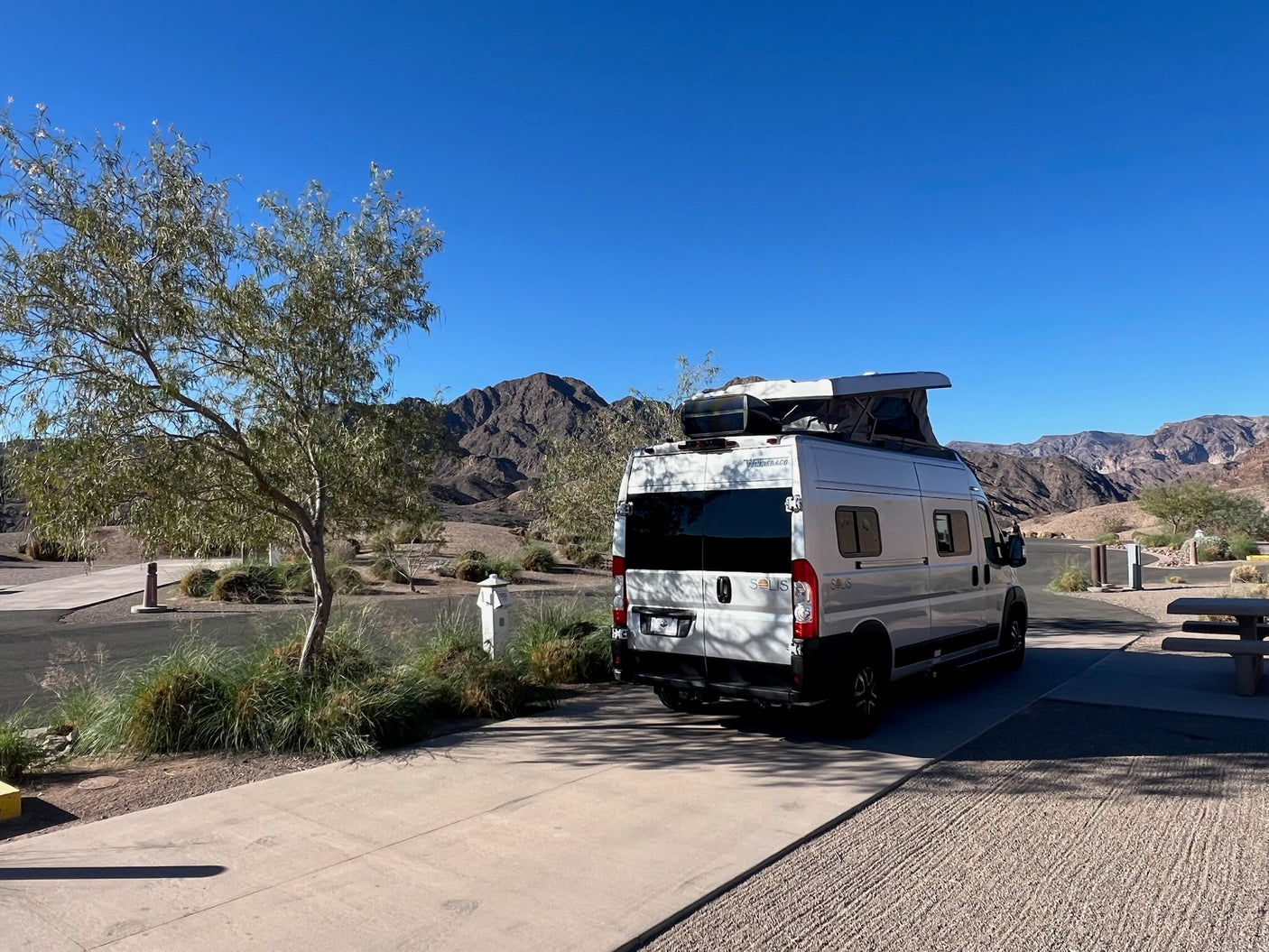 White Winnebago Solis rv rental camper van with pop-top extended at a desert campsite, mountains under a clear blue sky.