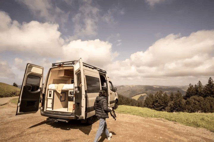 parked on a dirt path overlooking green hills.|Relax in your spacious RV rental! A woman enjoys the custom camper van's cozy interior with rear doors open to nature.|Your next rv rental adventure starts here! Camper van with open doors — photo 3