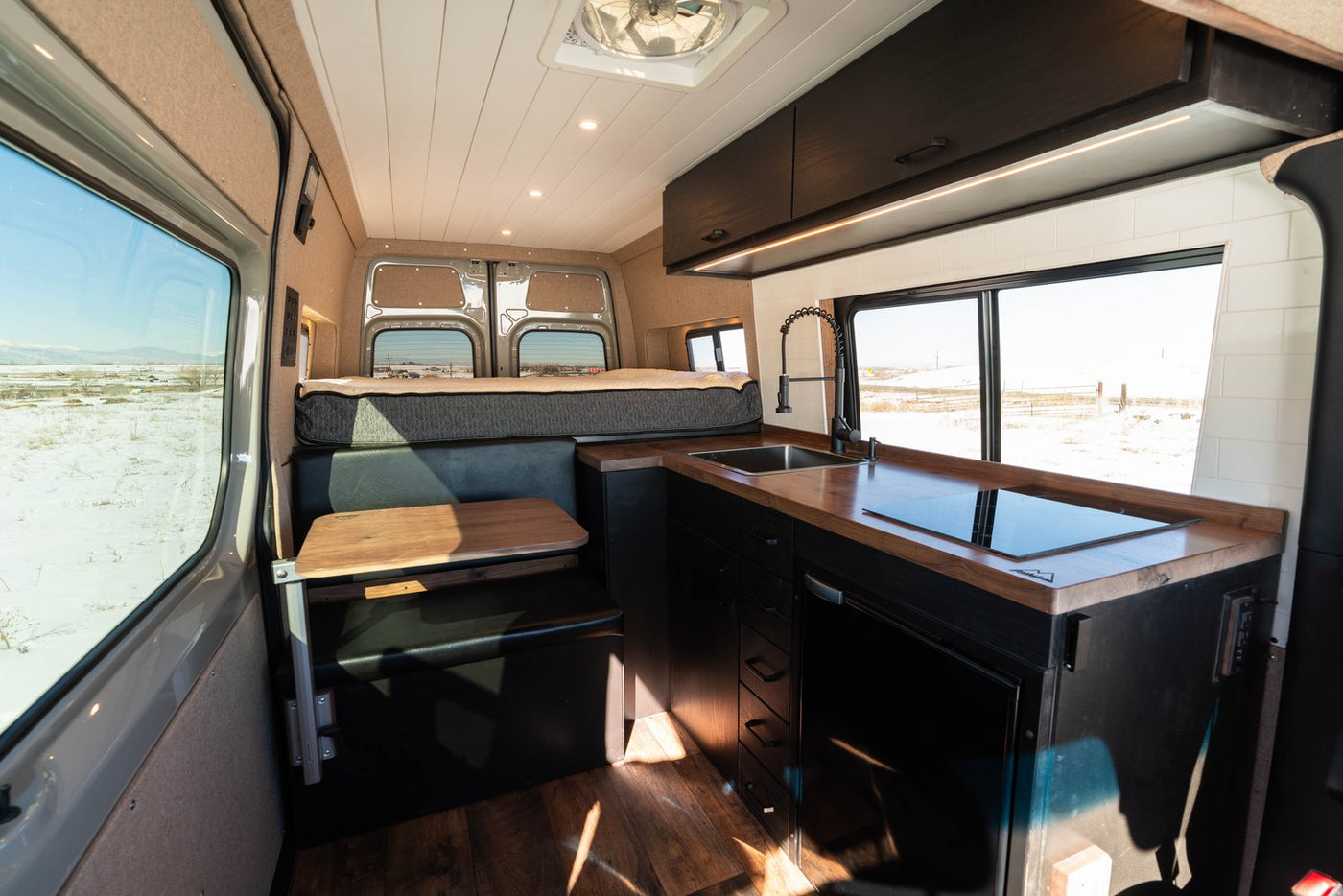 and a scenic snowy mountain view from the window. Perfect for your next adventure.|This RV rental's versatile interior features a lifted bench seat revealing ample storage and a compact fold-down table.|View inside an RV rental camper van: modern kitchen with dark cabinets — photo 10