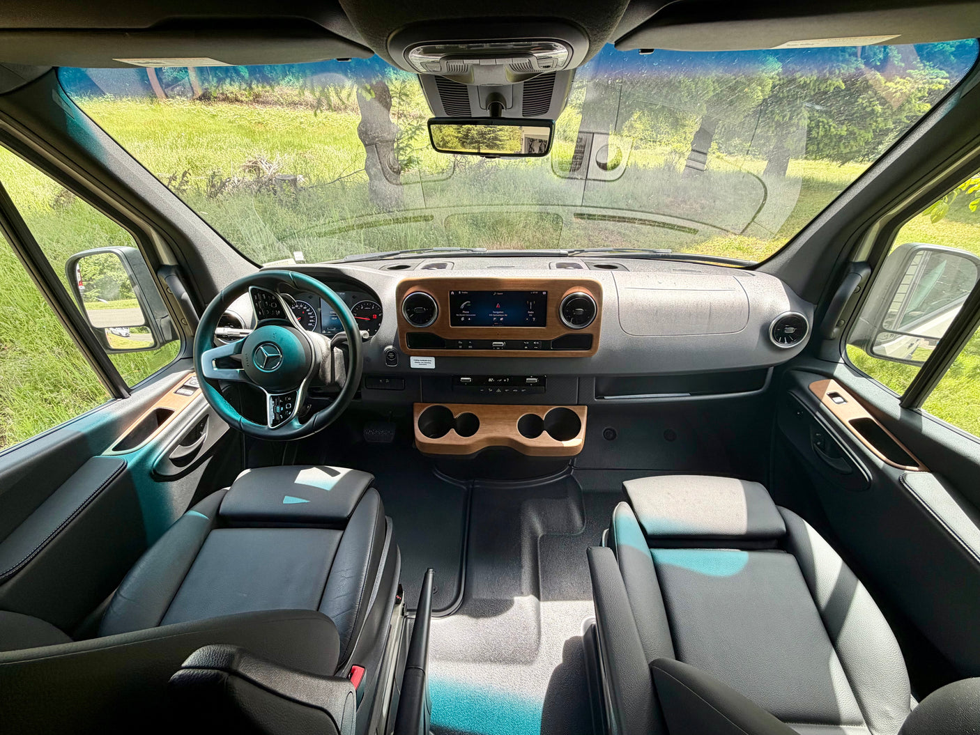 Front cabin of an RV rental with a modern dash, wood accents, comfy seats, and a clear view of green outdoors.