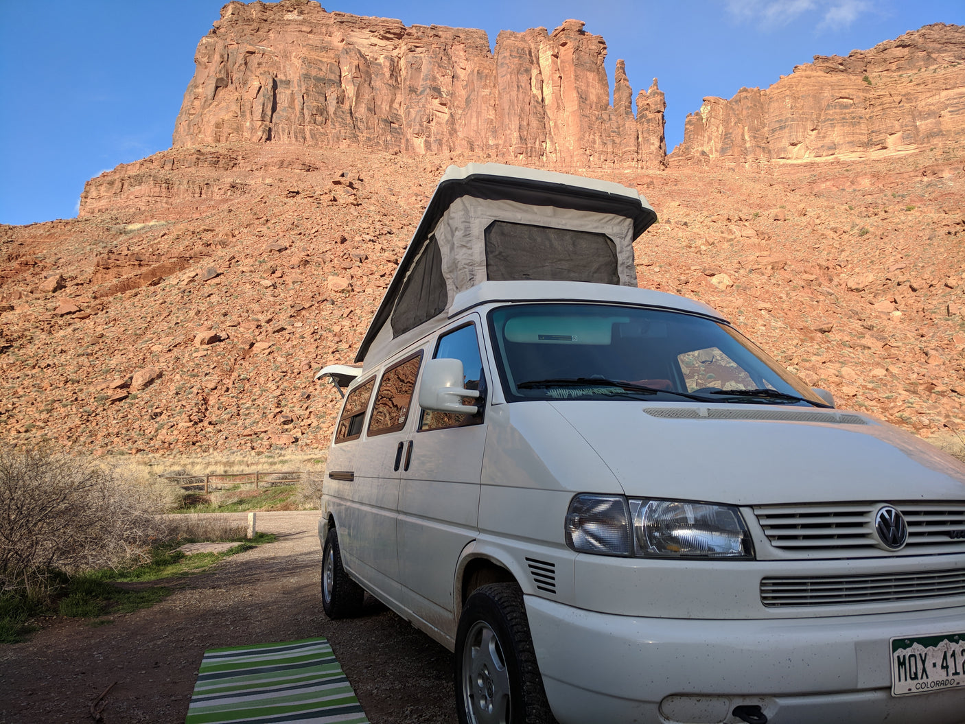An RV rental pop-top camper van faces towering red rock cliffs, parked on a dirt path with a striped mat. Ideal for desert adventures.