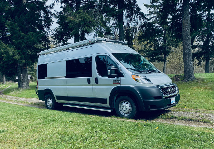 Silver Ram ProMaster RV rental, with an awning, parked on a gravel path by tall evergreen trees.