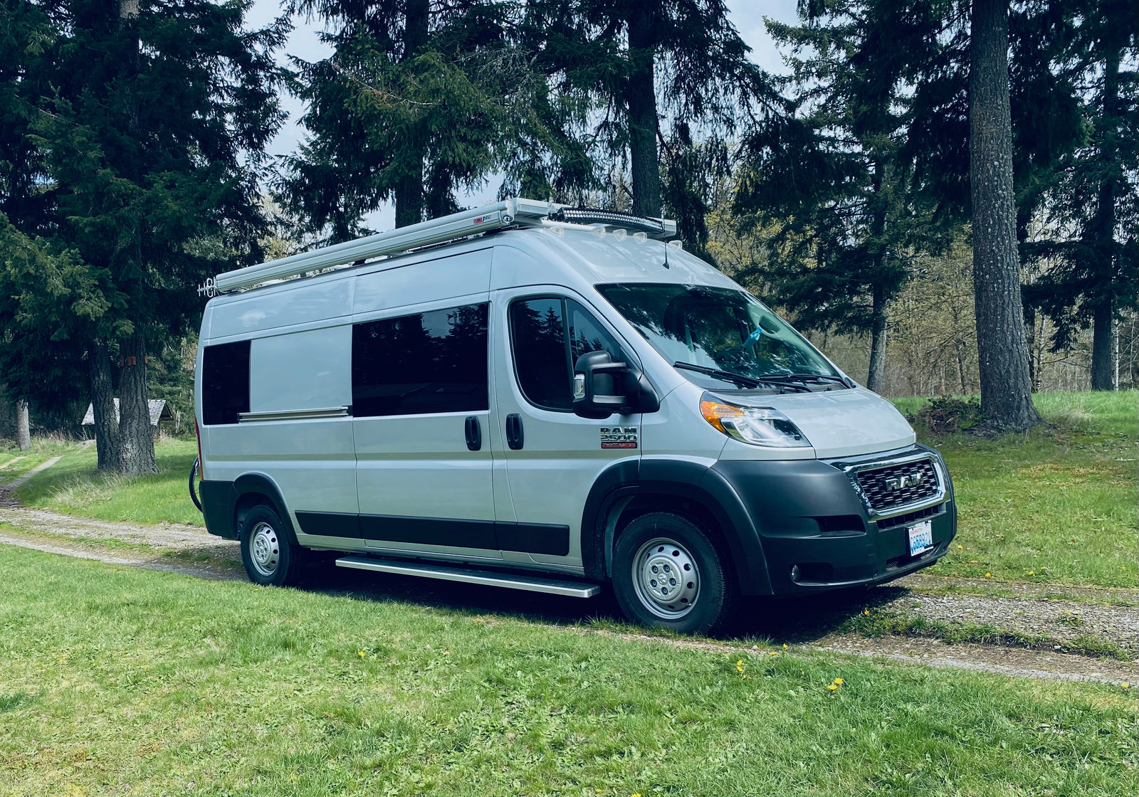 Silver Ram ProMaster RV rental, with an awning, parked on a gravel path by tall evergreen trees.