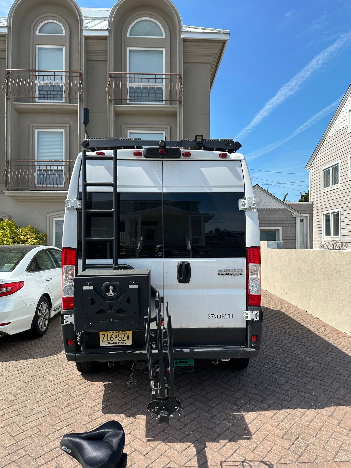 White Ram ProMaster RV rental van equipped with a ladder and bike rack, seen parked on a brick driveway.