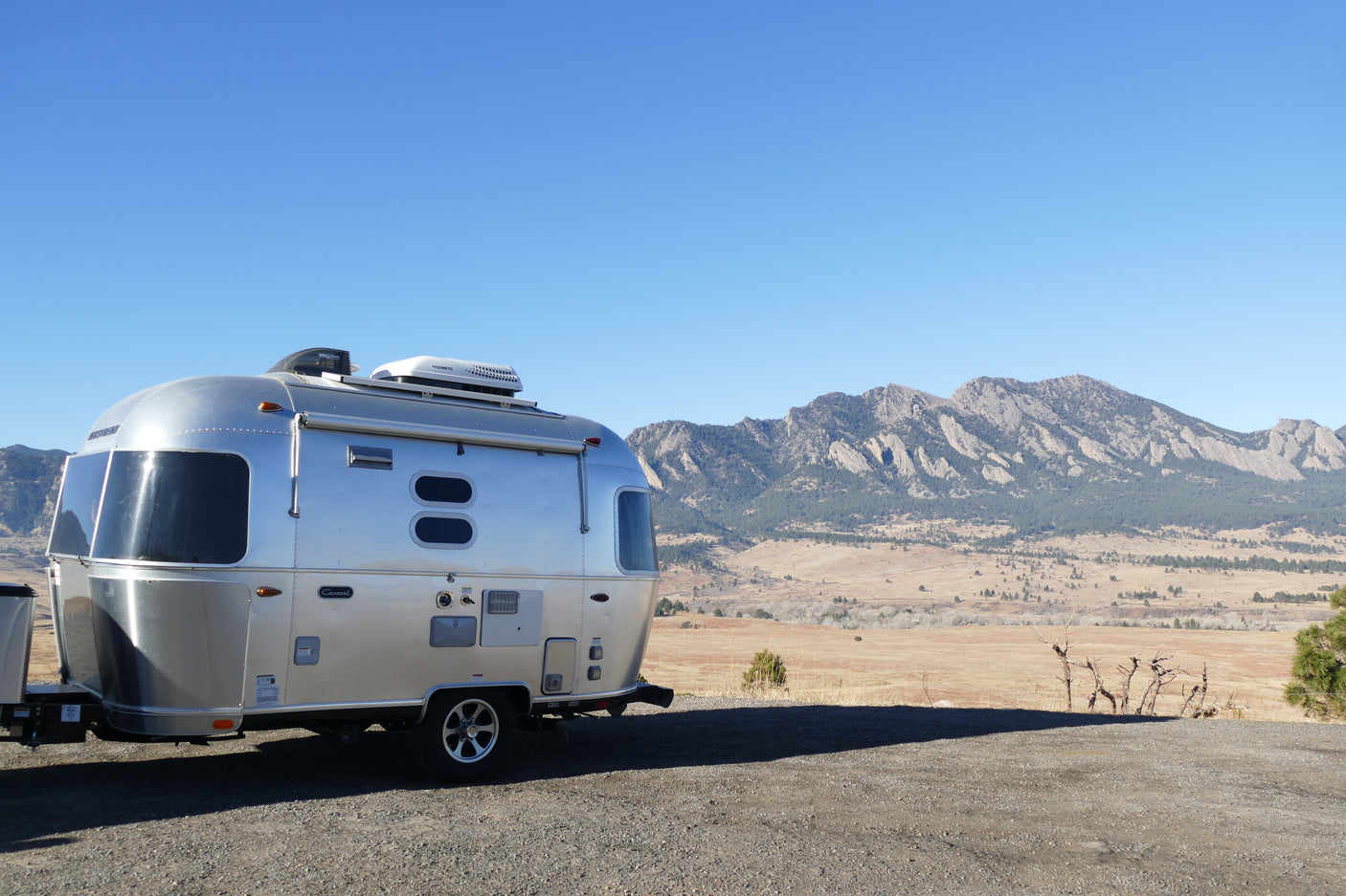 An Airstream RV rental is parked on gravel, facing majestic mountains and a sprawling plain under a bright blue sky.