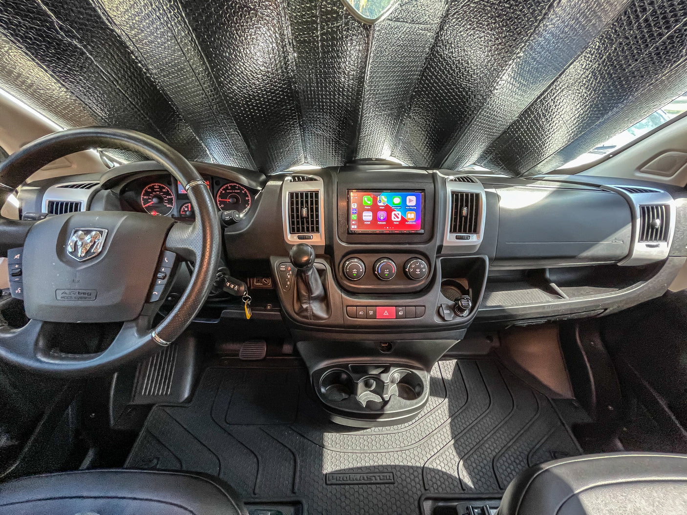 Inside a Ram ProMaster rv rental van: dashboard view with a sunshade, steering wheel, infotainment, and floor mats.
