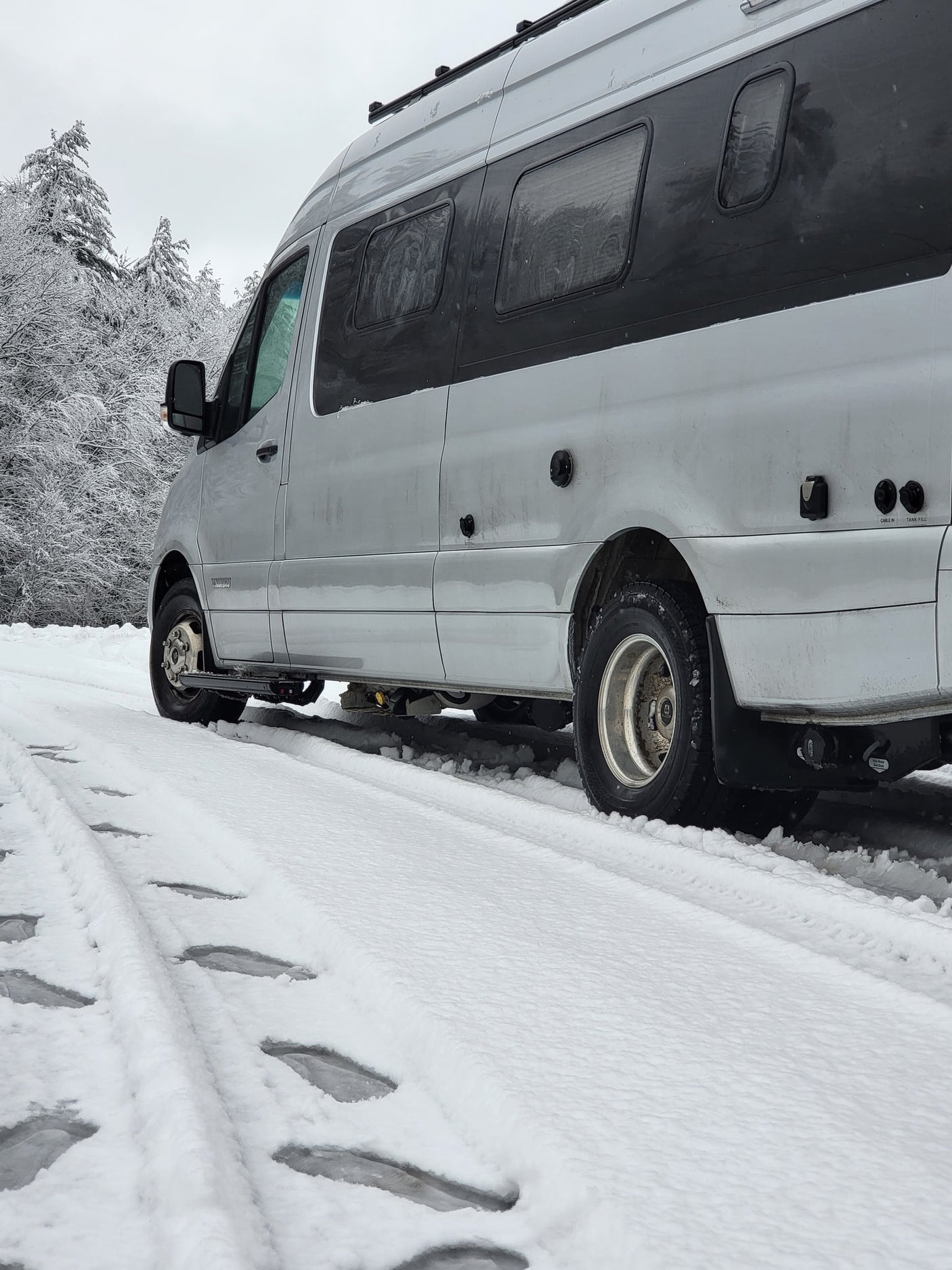 Silver camper van RV rental parked in deep snow. Tire tracks visible. Snowy trees in background, overcast sky.