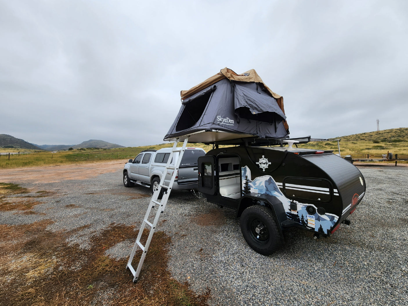 RV rental setup: Silver truck, black teardrop camper with SkyeDen rooftop tent, and ladder on gravel, ready for adventure.