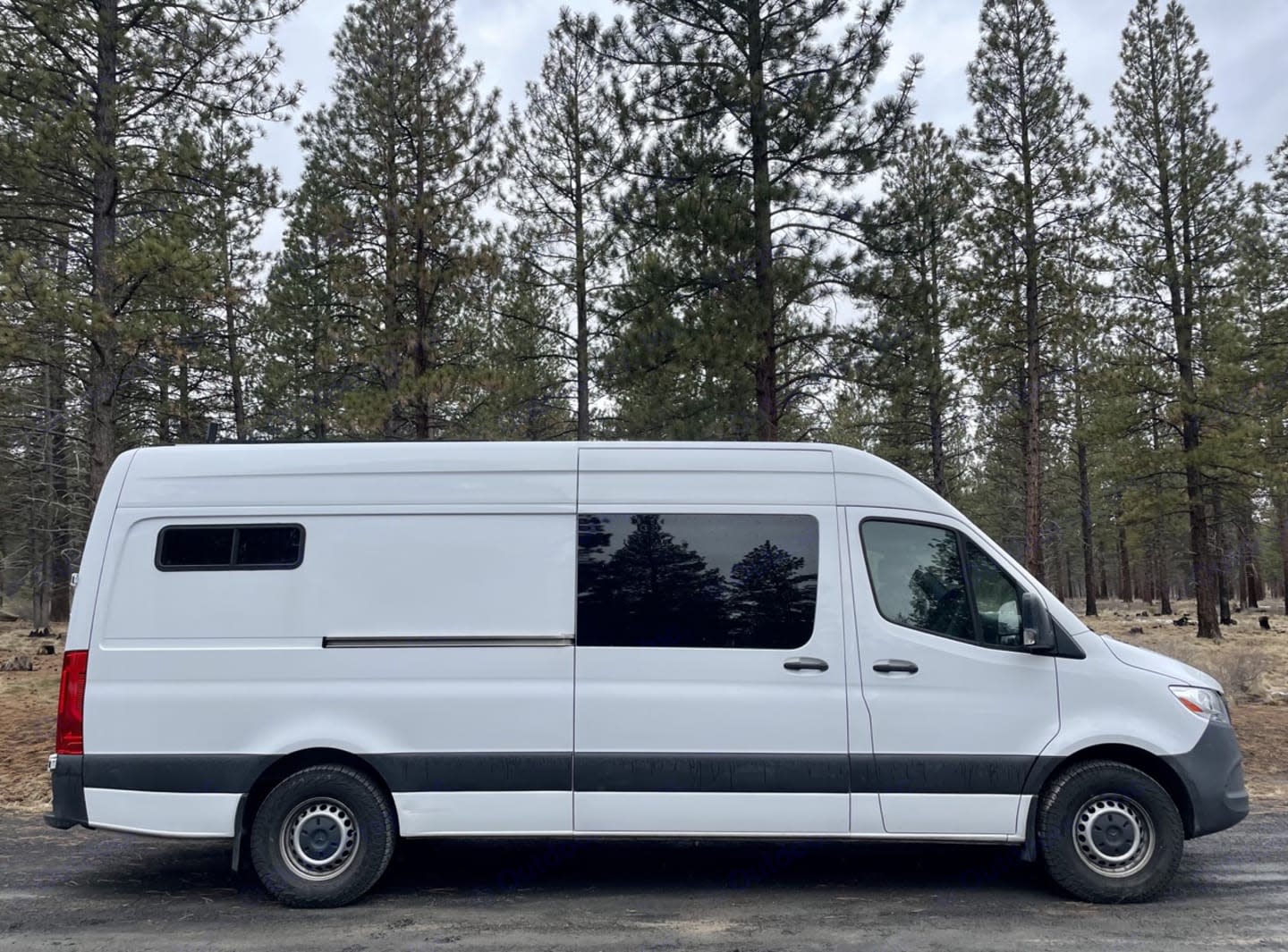 Side view of a white camper van, perfect for rv rental, parked on a dirt road by pine trees.