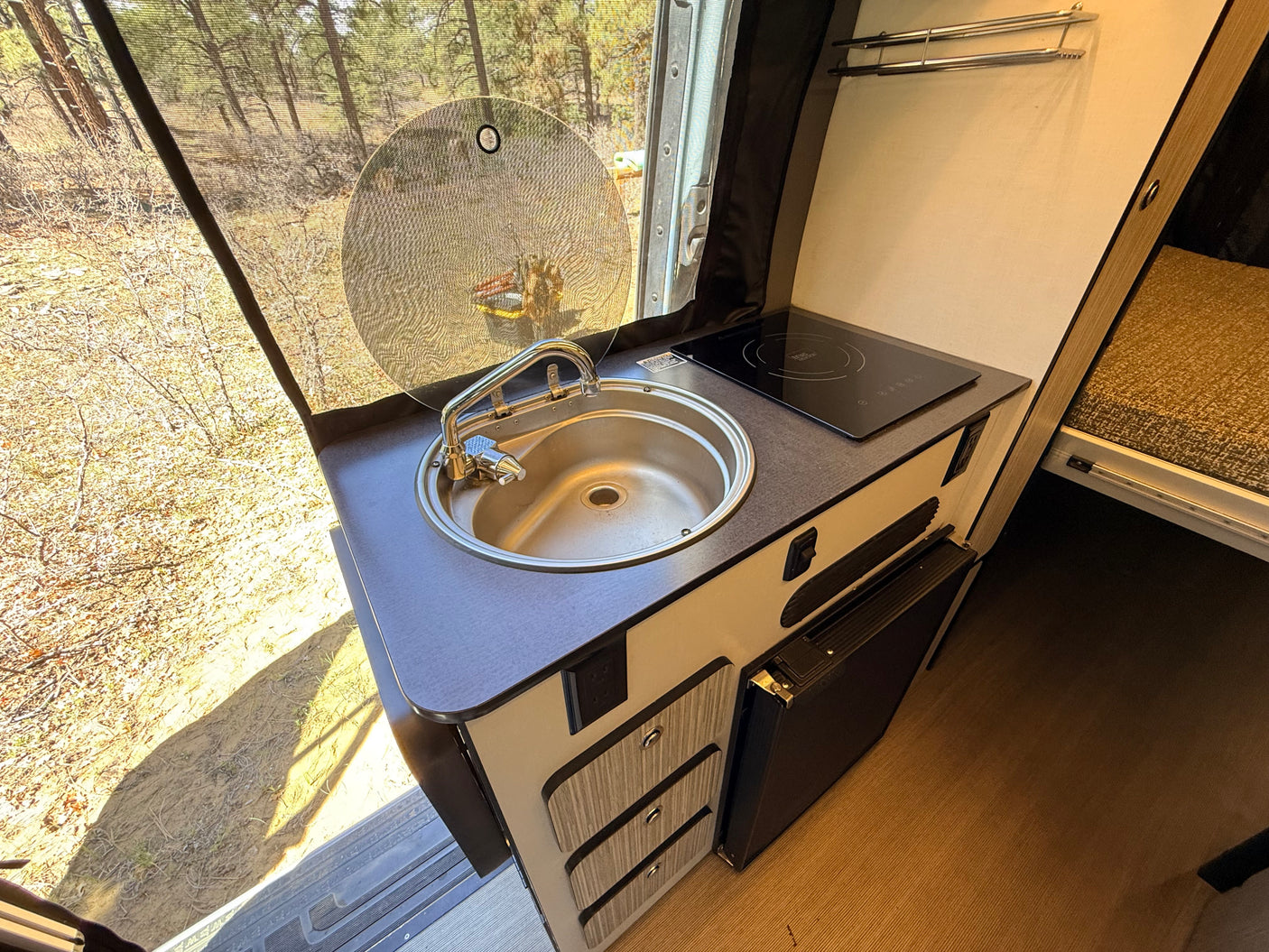 RV rental kitchen: compact layout with sink, induction cooktop, small fridge. Screened window offers a view of nature.