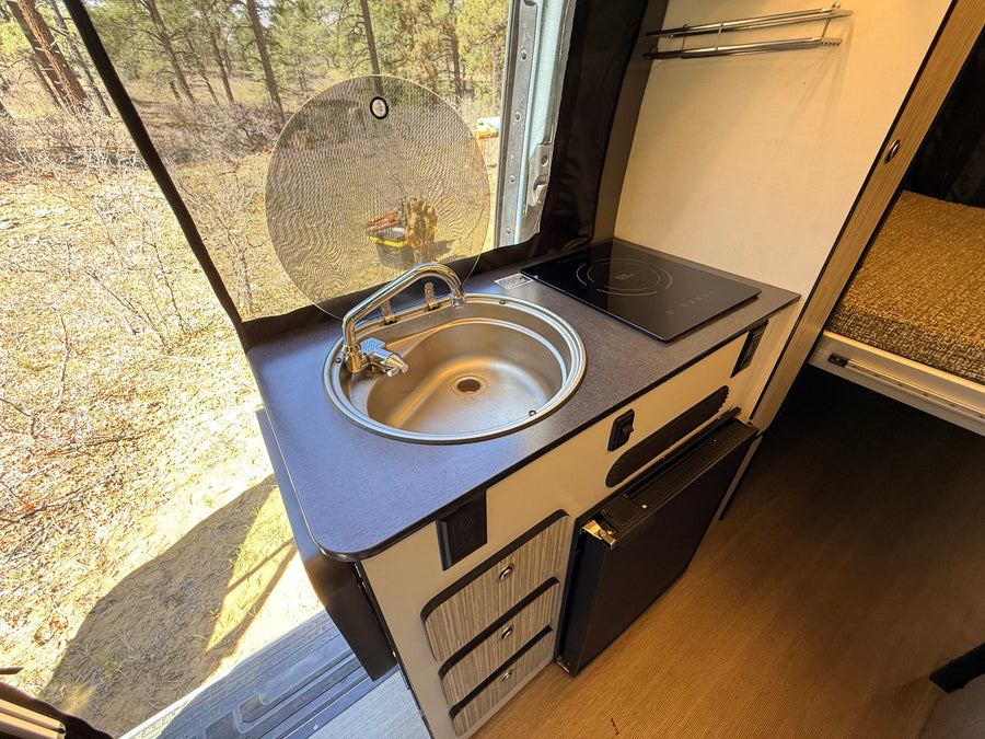 RV rental kitchen: compact layout with sink, induction cooktop, small fridge. Screened window offers a view of nature.