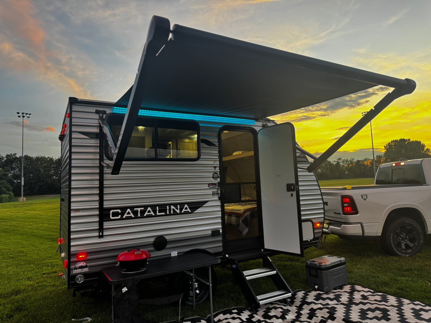 A Catalina travel trailer rv rental at sunset. Note the extended awning with blue lights, outdoor mat, grill, and truck in a grassy field.