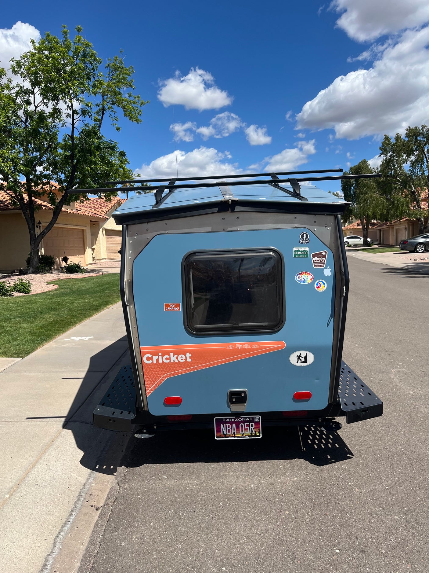 A light blue Cricket camper, a great option for an RV rental, is parked on a residential street, adorned with various travel stickers.