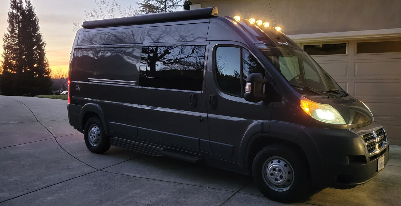 Dark gray camper van rv rental with lights on, parked on a driveway at sunset. Tree reflections in windows.