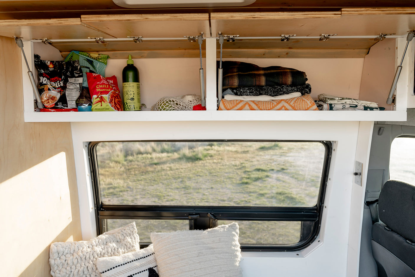 Inside an RV rental: organized overhead cabinets with snacks, cooking oil, and cozy blankets. A window reveals a grassy field.