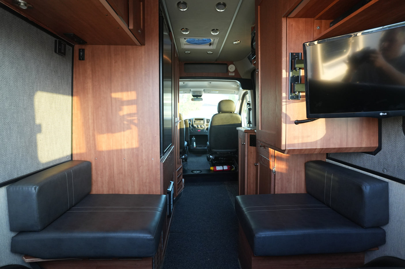 Interior of an rv rental: opposing dark benches, wood cabinetry, a TV, and the driver's cabin in view.