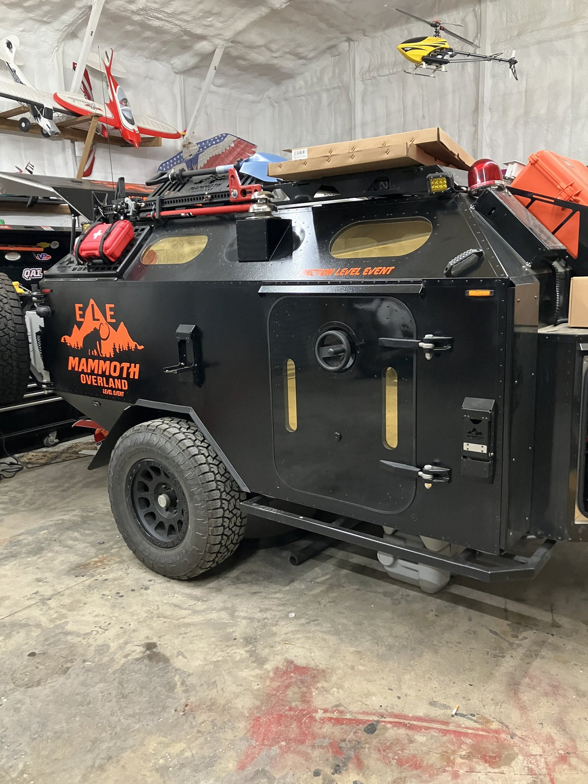 Side view of the 2023 Mammoth Overland ELE off-road trailer in a shop: side entry door with a round porthole window and two slit windows, orange MAMMOTH OVERLAND ELE graphics on the black body panel, roof rack loaded with recovery gear and a fuel can, and an all-terrain tire on a black wheel — photo 2