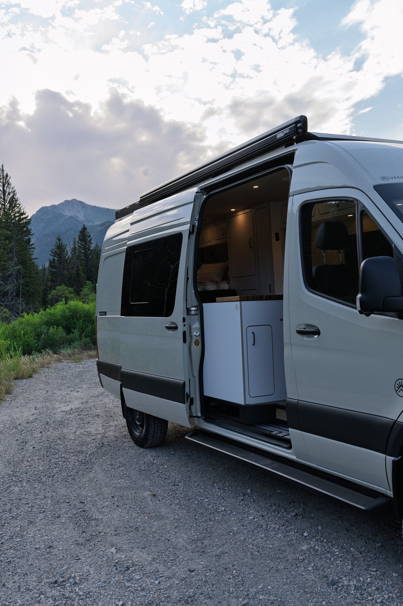 Light grey RV rental camper van parked on a gravel road with open side door, showing interior. Mountains and trees in background.