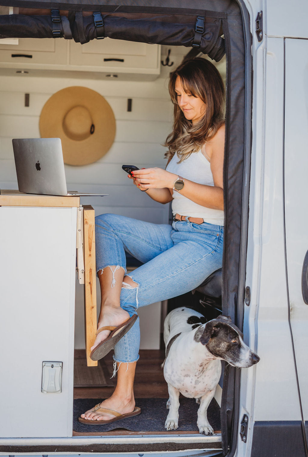 Woman sits in an RV rental with phone & laptop, dog nearby. Van life dream for remote workers.