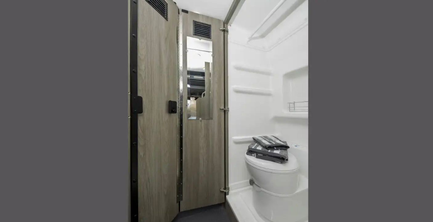 RV rental wet bath features a white toilet with towels, shower area, and a wood-look door with an integrated mirror.