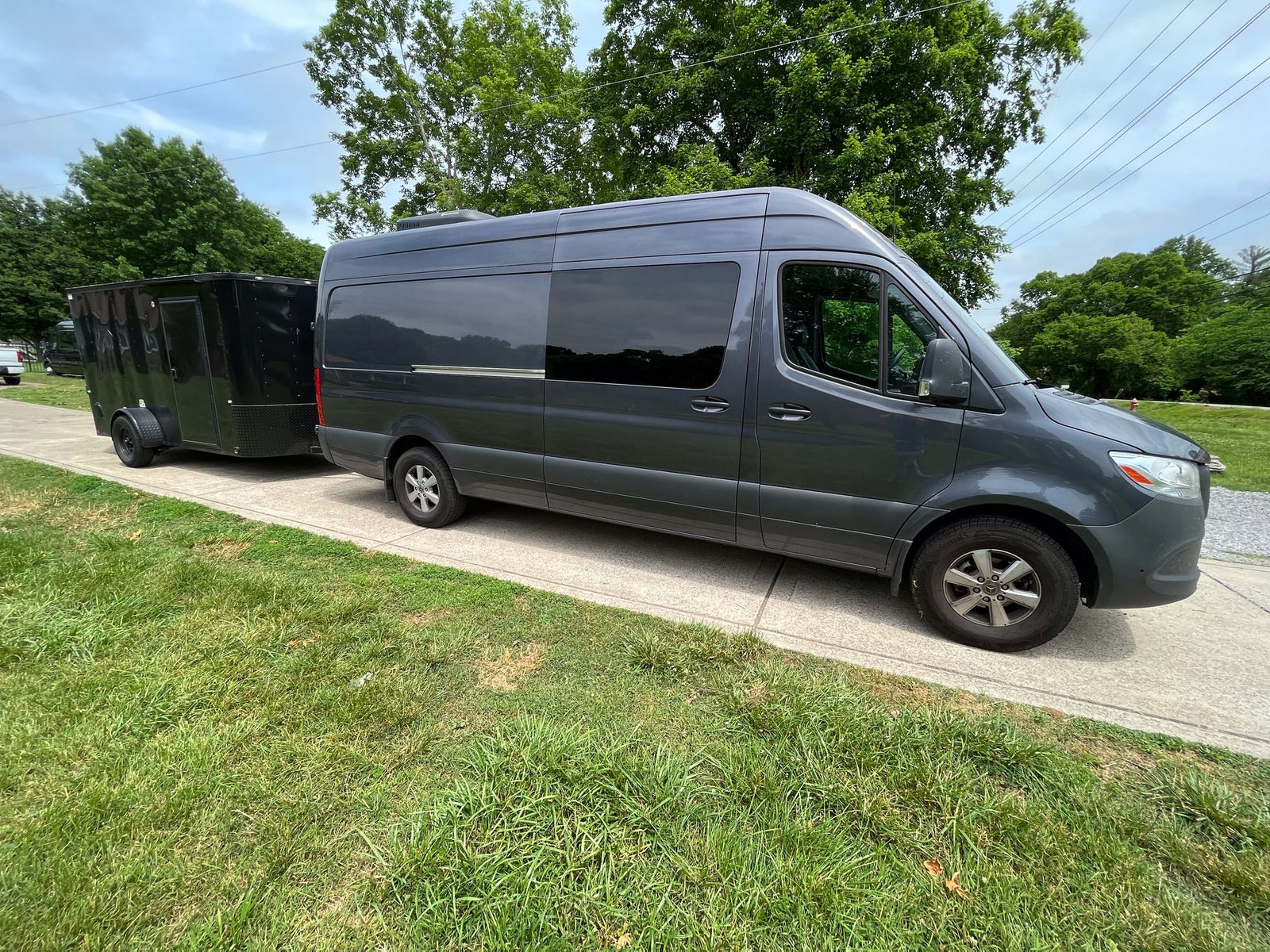 A grey RV rental Sprinter van towing a black trailer on a path beside green grass and trees.