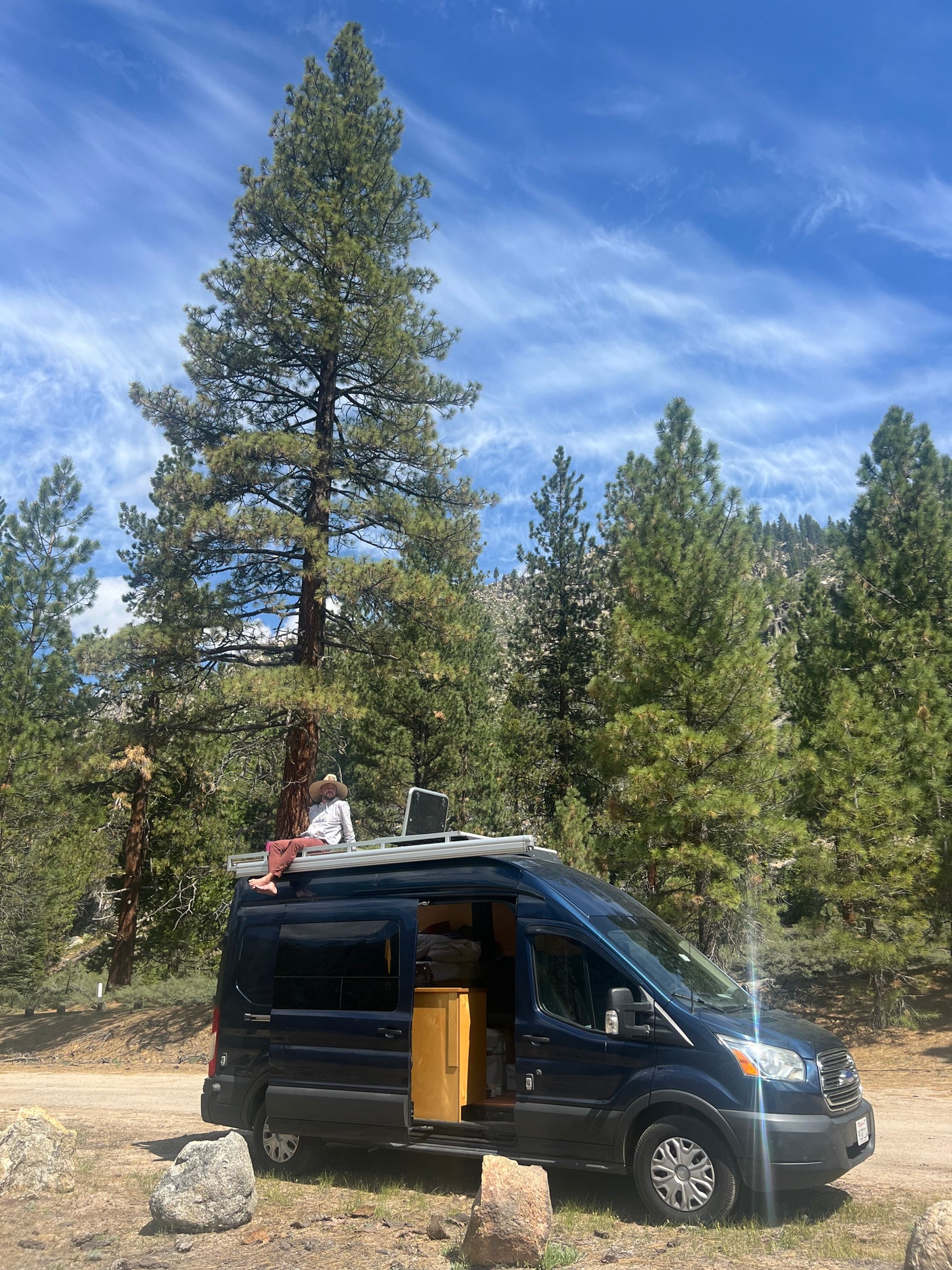 A person sits on a dark blue camper van's roof rack amidst tall pine trees under a blue sky. Experience an RV rental adventure!