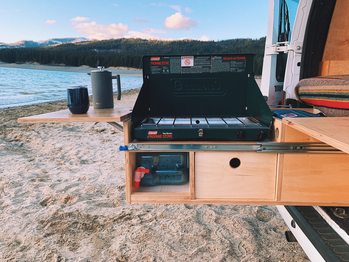 RV rental adventure! Lakeside outdoor kitchen setup with a Coleman stove, coffee, and water tap, perfect for van life camping.