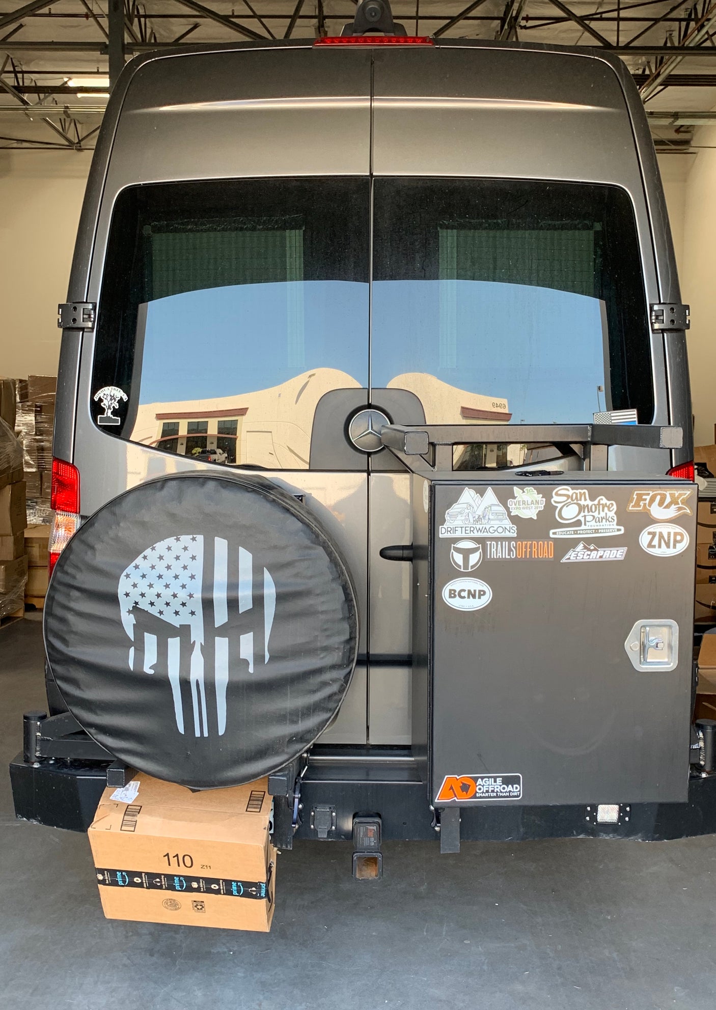 Rear view of a customized gray RV rental, featuring a skull tire cover, storage box with off-road stickers, and a Mercedes logo.