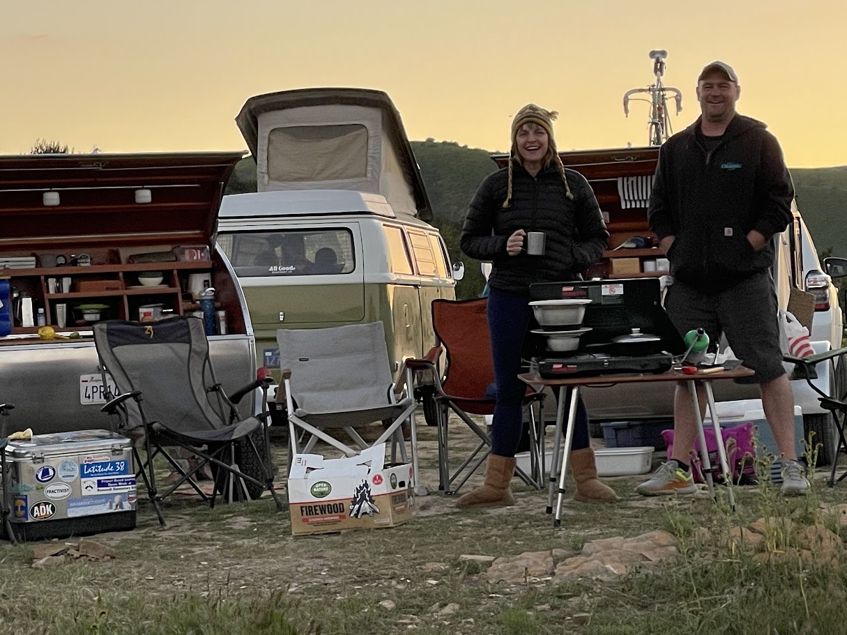 A couple smiles, cooking dinner with an RV rental campervan & truck setup, chairs, and cooler at dusk.