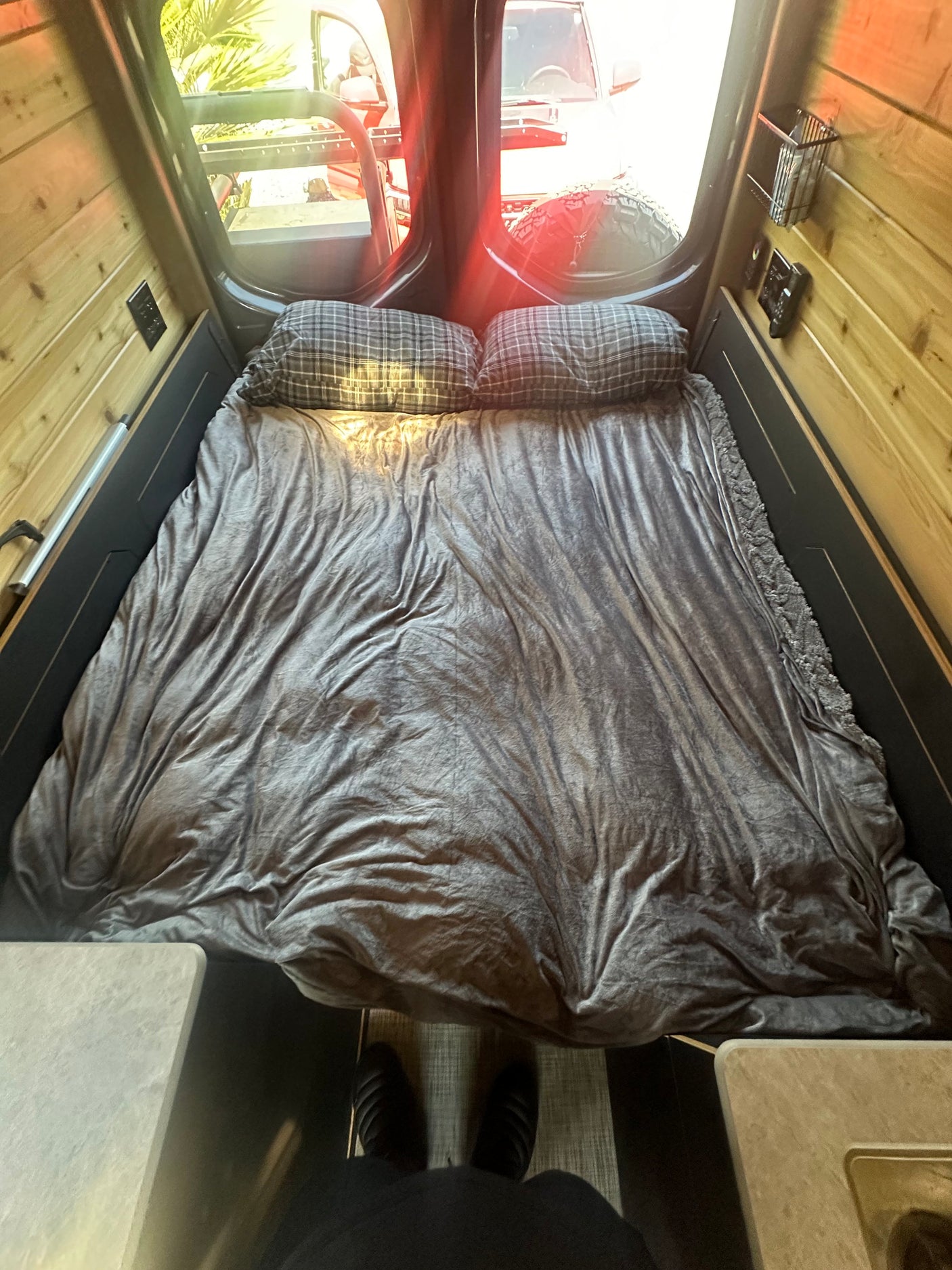Inside an RV rental: a neatly made bed with grey blankets and plaid pillows, surrounded by warm wood paneling. Rear window views.