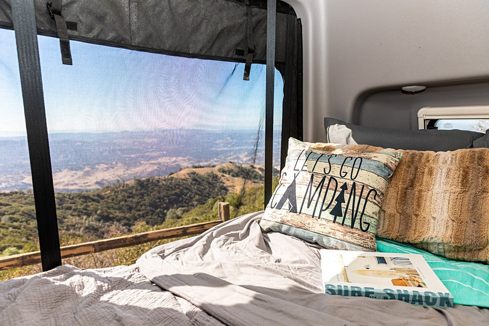 An RV rental bed, with a 'LET'S GO CAMPING' pillow, offers sweeping mountain views through a mesh screen.