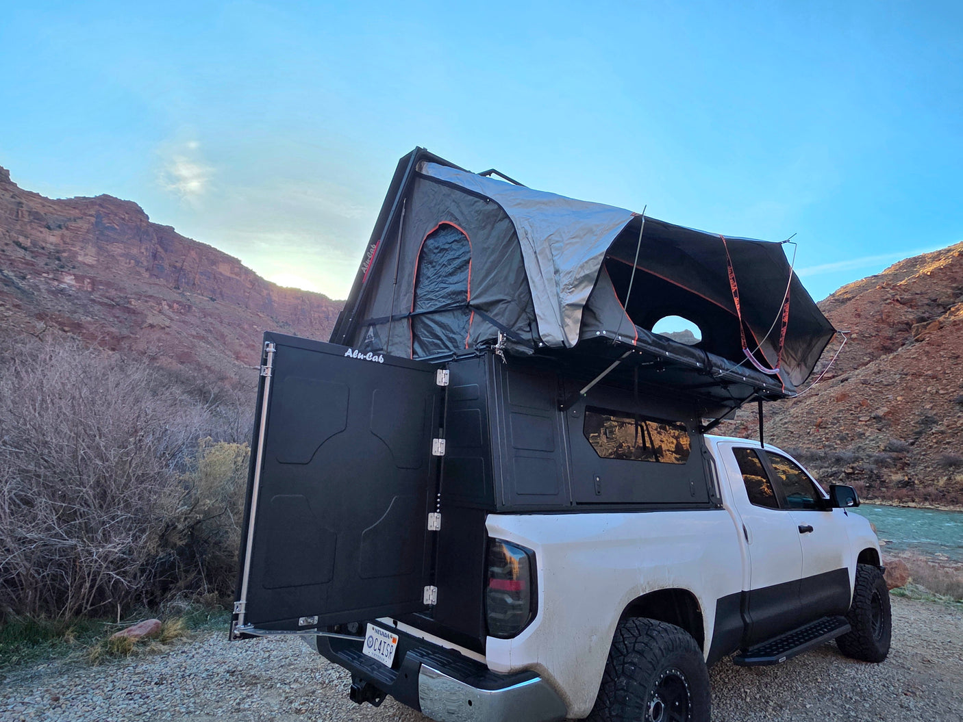 White truck with Alu-Cab camper and open rooftop tent as an RV rental, parked by a scenic river and rocky mountains.