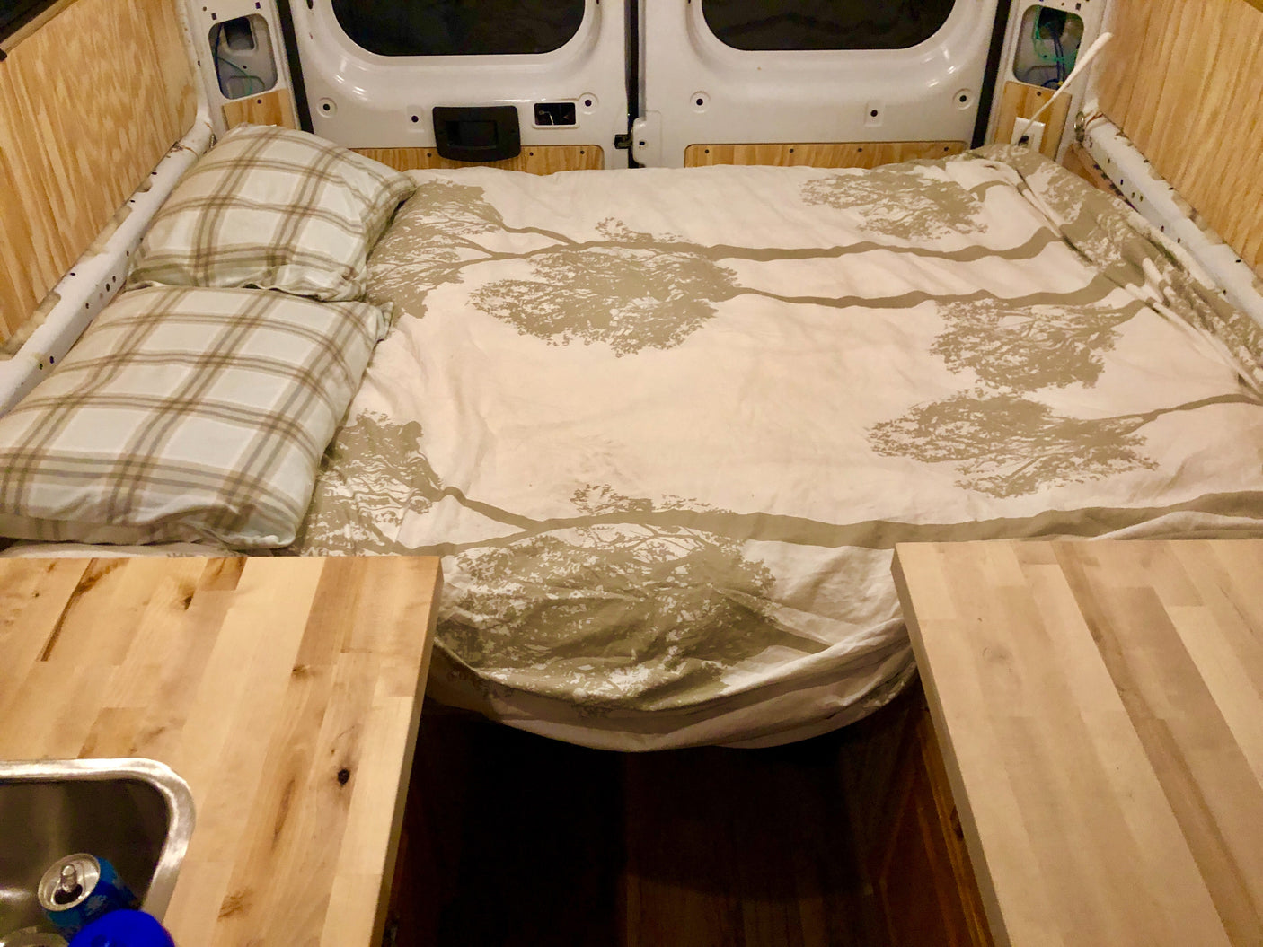 Inside a custom RV rental: a cozy bed with nature-themed bedding, surrounded by wood paneling, next to a mini kitchen.