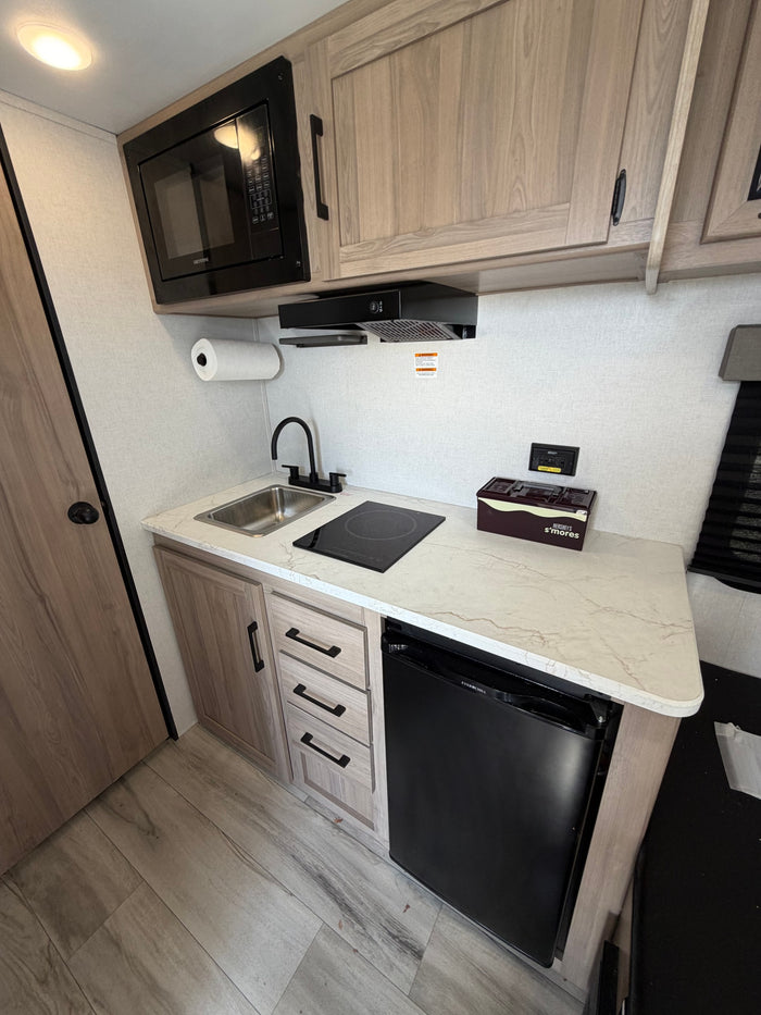 Modern RV rental kitchen with black appliances, including microwave, sink, induction cooktop, fridge, and light wood cabinetry.