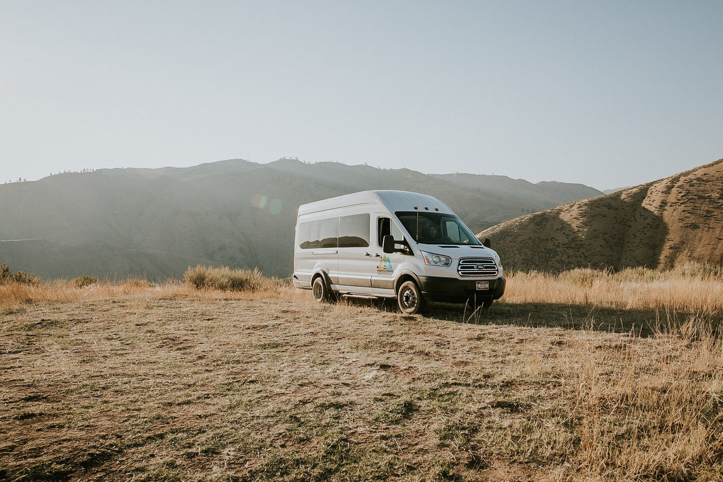 A white RV rental van is parked on a dry, grassy hillside with scenic mountains and a clear sky in the background.