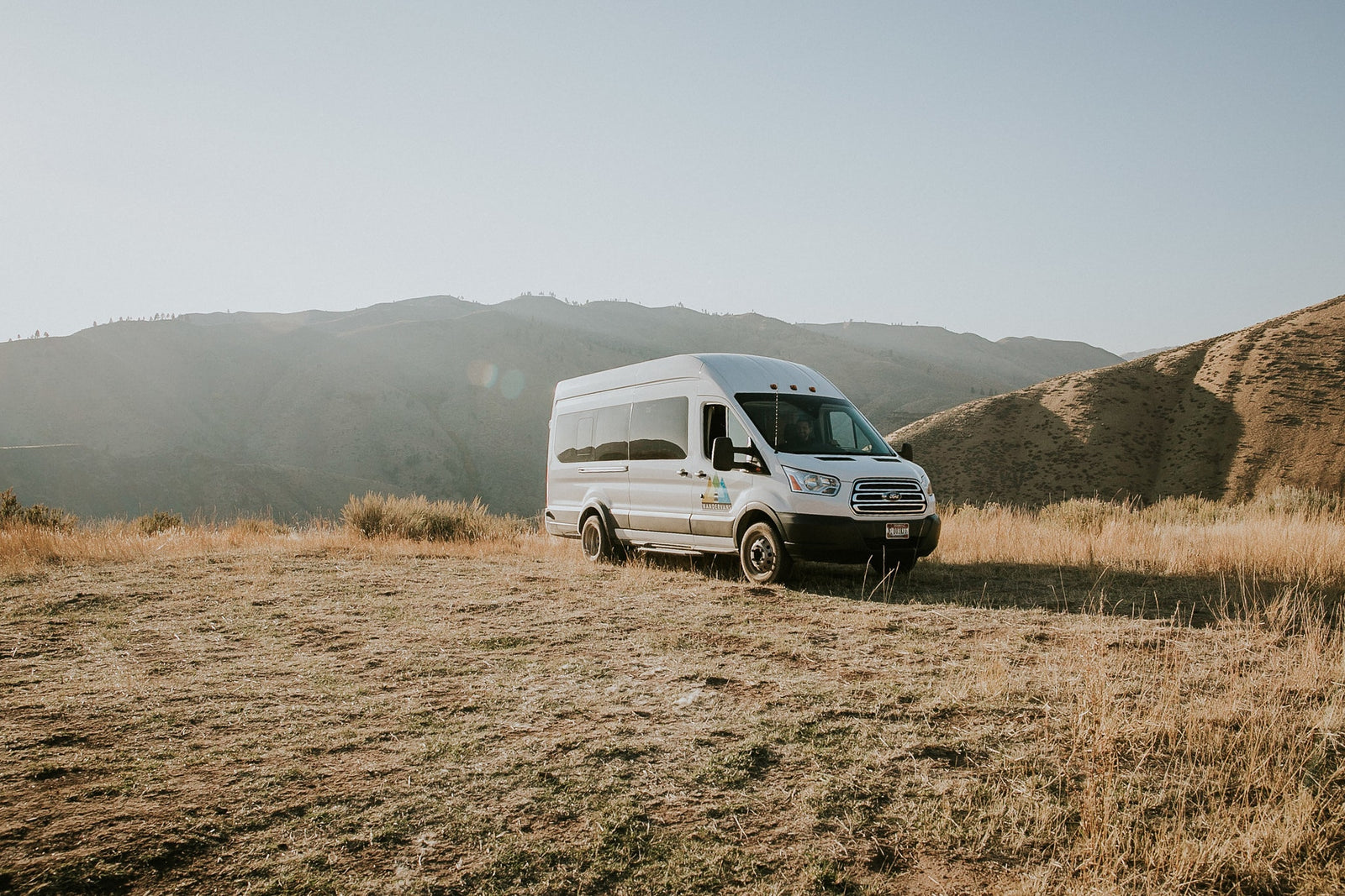 A white RV rental van is parked on a dry, grassy hillside with scenic mountains and a clear sky in the background.