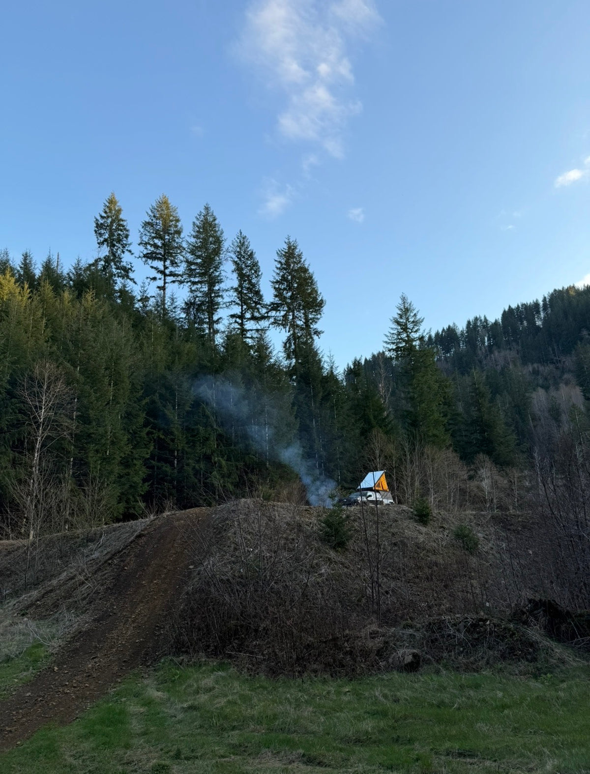 An RV rental with a pop-up tent atop a car, nestled in a forest clearing with a smoking campfire under blue skies. — photo 5