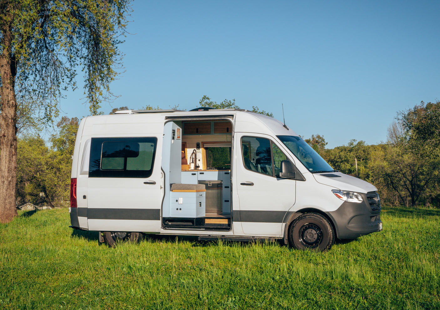 White RV rental camper van parked in a sunny field, side door open revealing a modern van life kitchen for adventure.