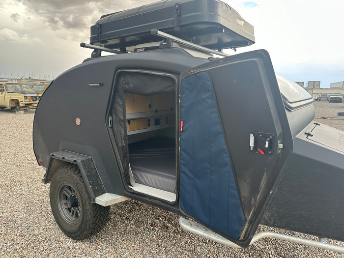 Black off-road teardrop camper RV rental with open door reveals interior bed, storage, and rooftop tent.