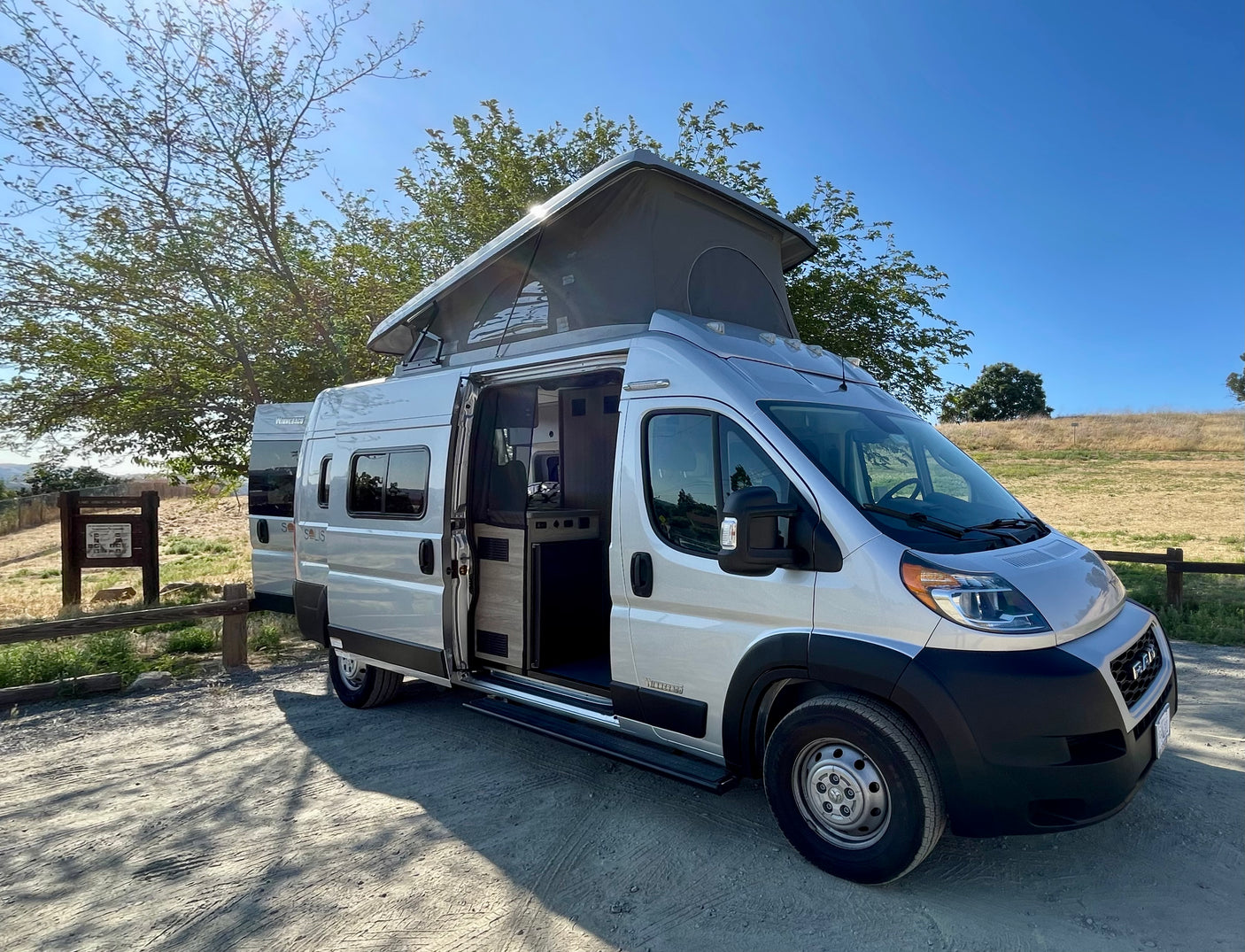 Silver pop-top RV rental, its side door open showing the interior, parked on a sunny dirt road.
